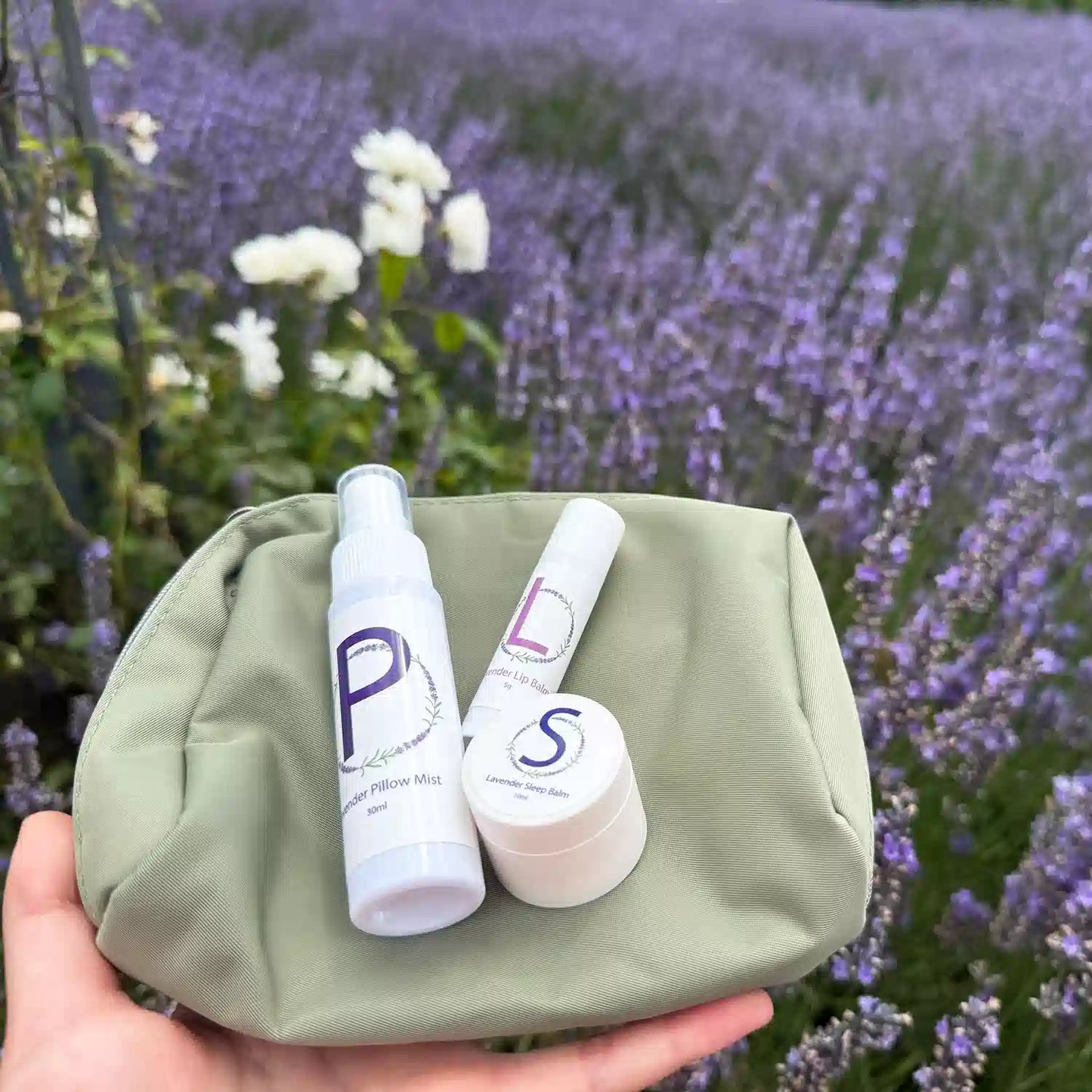 Easy Sleep Lavender Travel Kit, including a Travel-Sized Lavender Pillow Mist, Travel-Sized Sleep Balm, Travel-Sized Lavender Lip Balm, and an Earthy Green Toiletry Bag, beautifully arranged in a lavender field with white roses in the background.