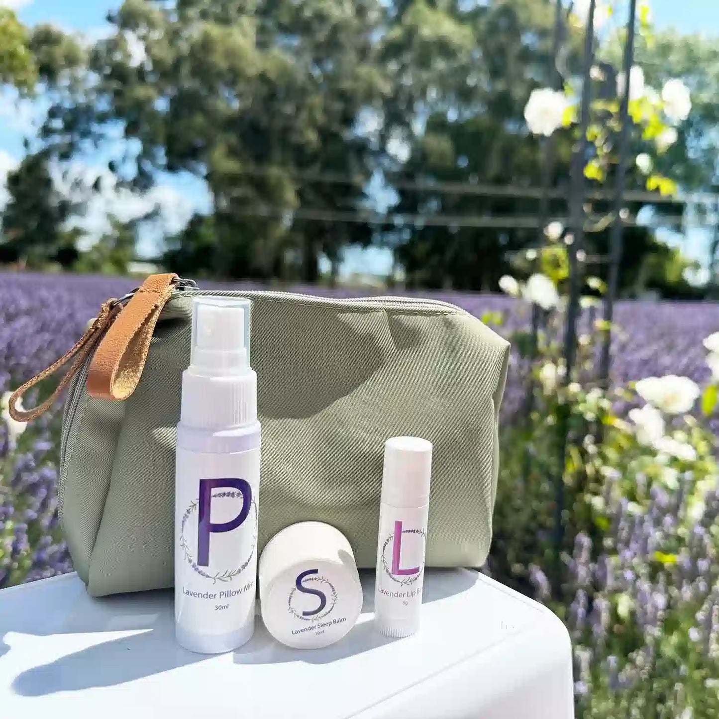 Lavender Easy Sleep Travel Essentials, featuring a Travel-Sized Lavender Pillow Mist, Travel-Sized Sleep Balm, Travel-Sized Lavender Lip Balm, and an Earthy Green Toiletry Bag, displayed among a lavender field alongside white roses.