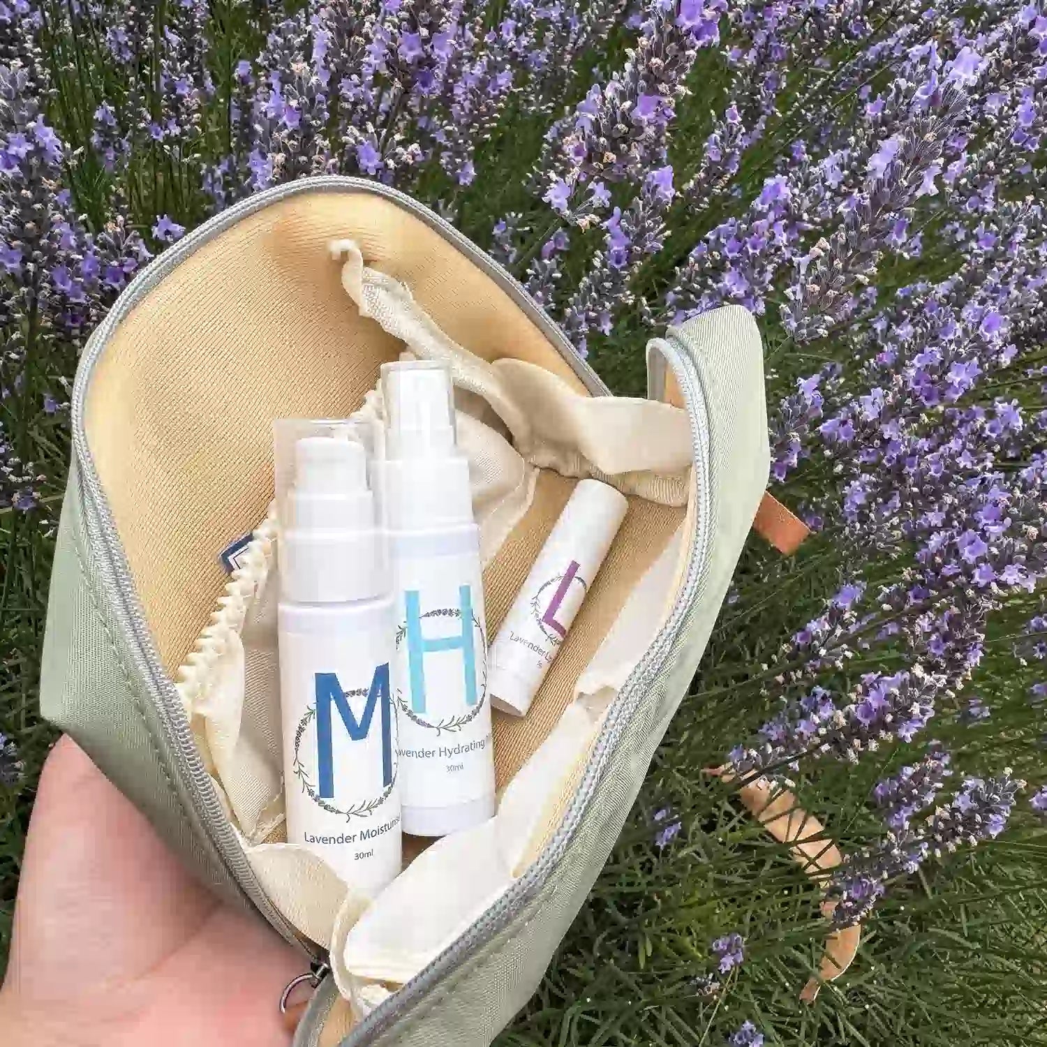 Lavender-Travel-Toiletries-Skincare-Kit, featuring a Travel-Sized Lavender Face Toner, Travel-Sized Moisturiser Lotion, Travel-Sized Lavender Lip Balm, and an Earthy Green Toiletry Bag, displayed among lavender flowers at Lavender Backyard Garden, NZ.