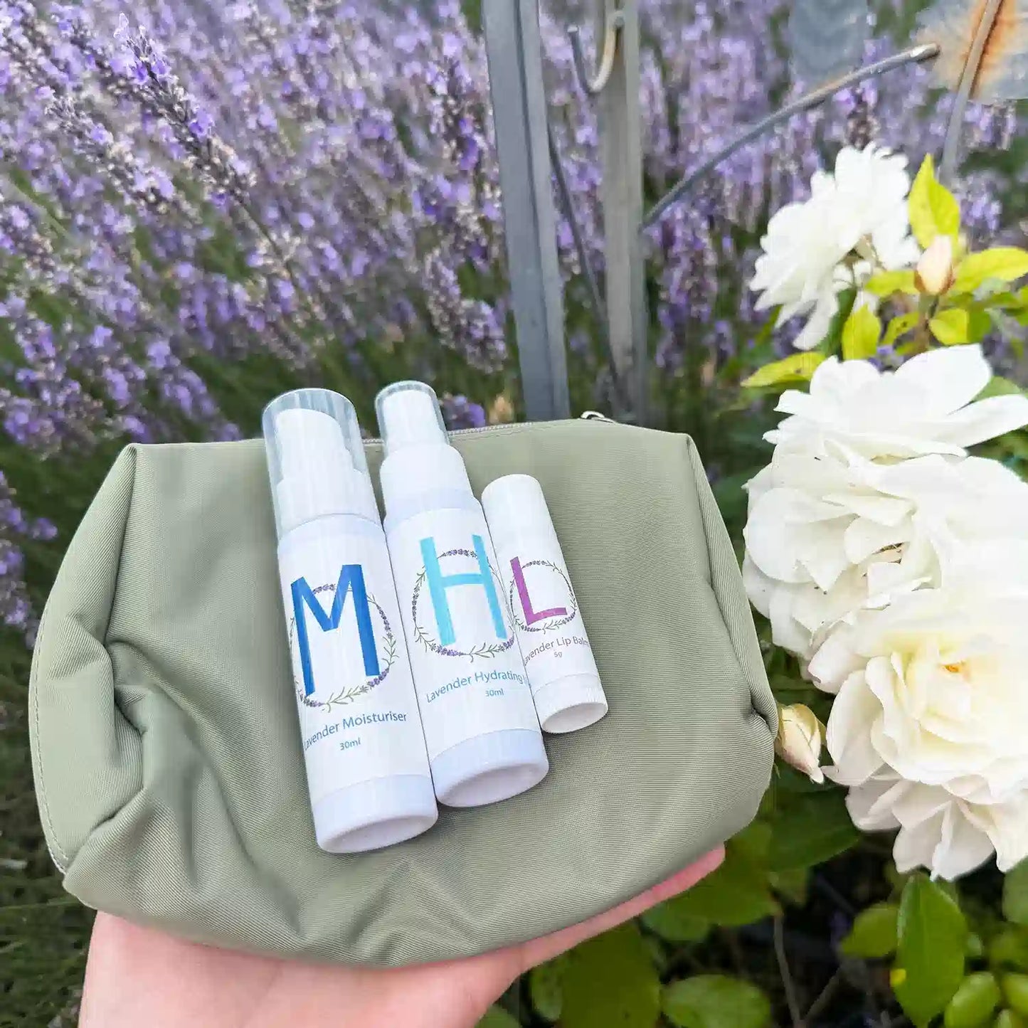 Lavender Travel Toiletries Skincare Kit, featuring a Travel-Sized Lavender Face Toner, Travel-Sized Moisturiser Lotion, Travel-Sized Lavender Lip Balm, and an Earthy Green Toiletry Bag, displayed against a vibrant lavender field at Lavender Backyard Garden, NZ, with delicate white roses alongside.