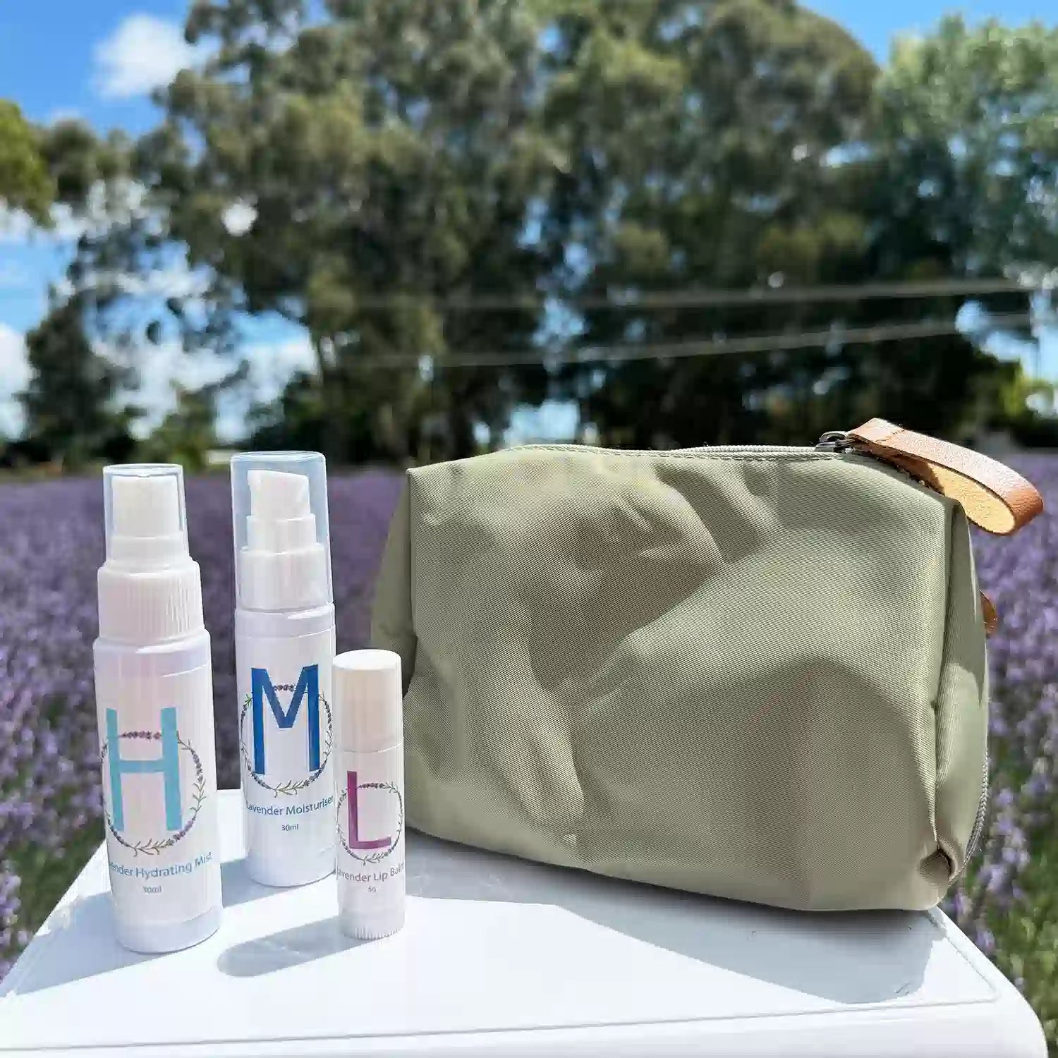 Lavender Travel Toiletries Skincare Kit, featuring a Travel-Sized Lavender Face Toner, Travel-Sized Moisturiser Lotion, Travel-Sized Lavender Lip Balm, and an Earthy Green Toiletry Bag, displayed among a lavender field with white roses in the background.