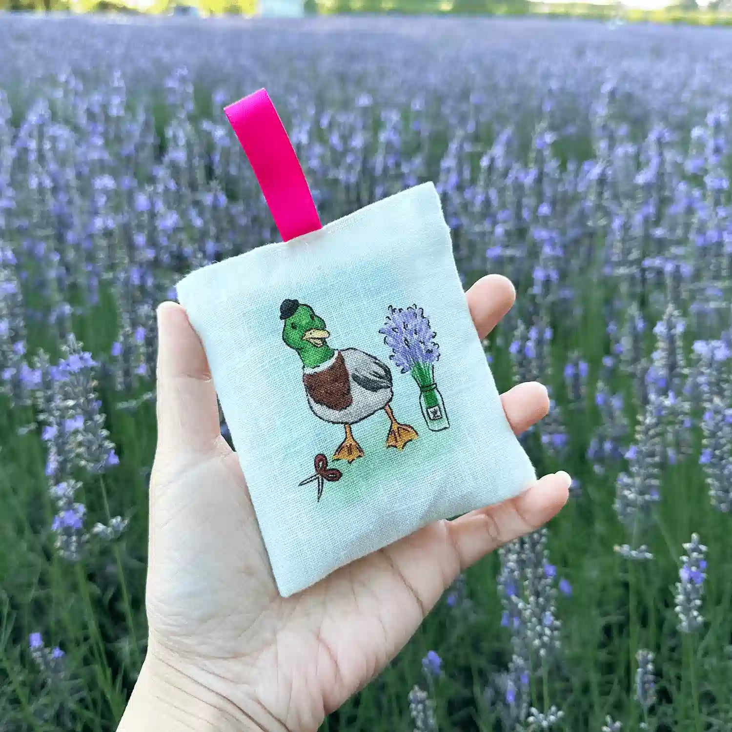 Duck of Avery, NZ-designed 100% linen sachets scented with lavender for closet or wardrobe, held by hand against a backdrop of lavender field at Lavender Backyard Garden NZ.