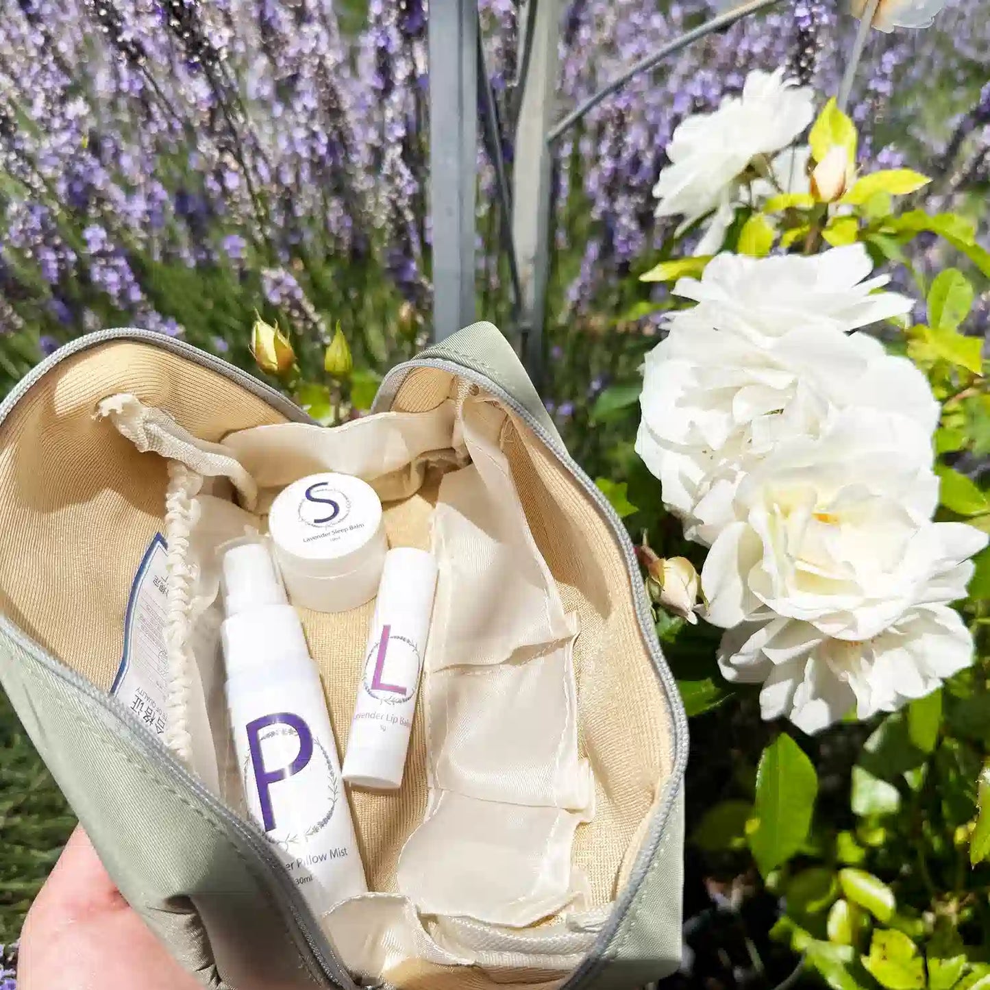 Sleep Well Travel Pack, including a Travel-Sized Lavender Pillow Mist, Travel-Sized Sleep Balm, Travel-Sized Lavender Lip Balm, and an Earthy Green Toiletry Bag, displayed in a serene lavender field with white roses in the background.