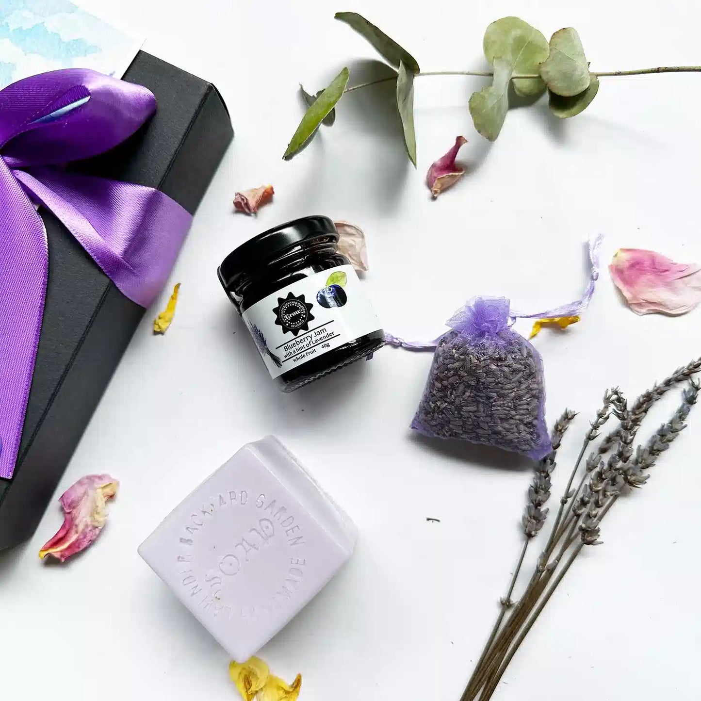 Close-up view of an "Affordable yet Thoughtful Gift Set," featuring a black gift box with a brown ribbon. Showcased items include a Dried Lavender Sachet, Blueberry Jam with a hint of Lavender (40g), and Lavender Goat Milk Handmade Soap, surrounded by mini dried lavender flowers, rosemary, and rose petals as background decor.