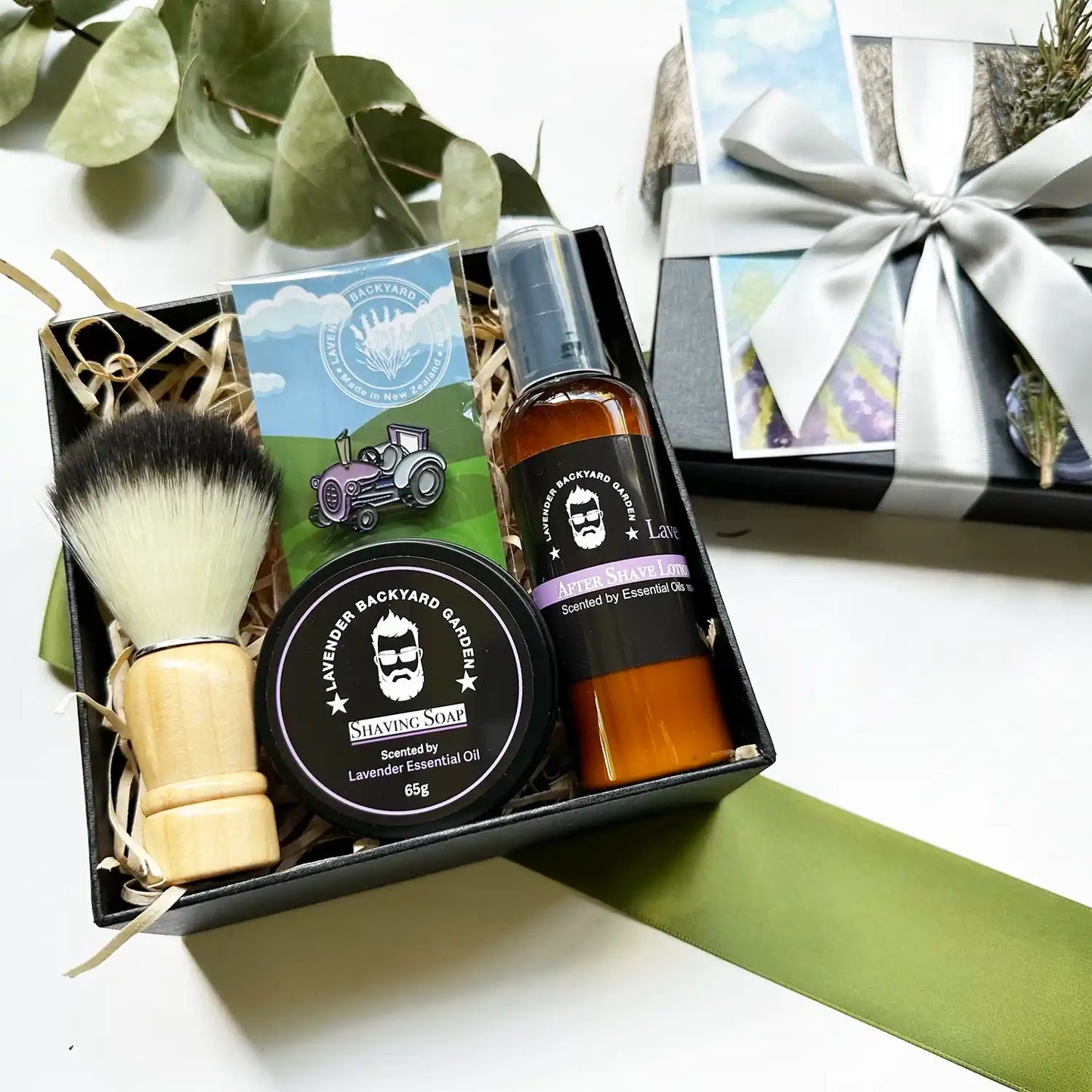 A beautifully arranged gift box featuring Lavender Soothing Shaving Soap, Lavender Men's Aftershave Refreshing Lotion, a Shaving Brush, and a Purple Tractor Badge, all elegantly wrapped with a silver ribbon and accompanied by a lavender field bookmark.