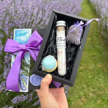 Close up view of Amazing Bath & Spa Giftset for Her small gift set scented by lavender essential oils, featuring Floral Bath Salts Tube, Dried Lavender Sachet, Lavender Essential Oil Bath Bomb (40mm), and Lavender Field Bookmark, against a backdrop of purple blooming lavender field.