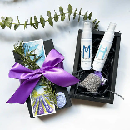 ravel gift set featuring lavender moisturizer and facial mist, lavender sachet, packaged in a black gift box with a purple ribbon, and a rosemary and lavender field bookmark.