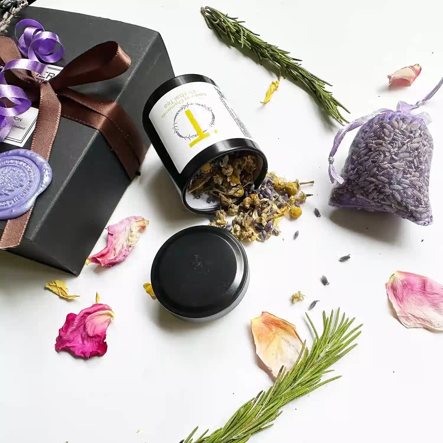 Close-up view of "Budget-Friendly Teacher Gift Ideas" set, showcasing a black gift box with a brown ribbon. Featured items include Lavender & Camomile Herbal Tea (Travel Mini) and a Dried Lavender Sachet, elegantly arranged with mini dried lavender flowers, rosemary, and rose petals as background decor.