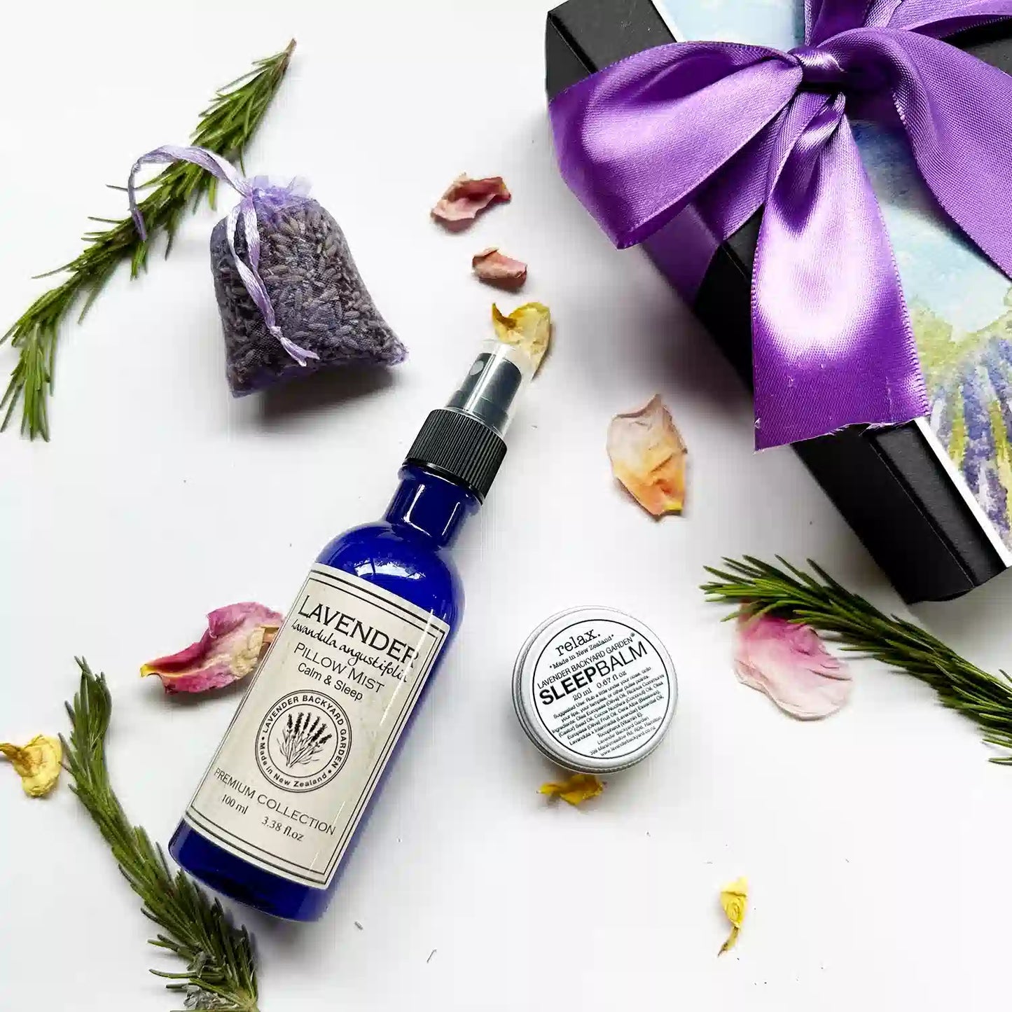 Close-up of "Lavender Sleep Balm, Lavender Sleepwell Pillow Mist, Dried Lavender Sachet" with background decoration of eucalyptus leaves and rose petals.