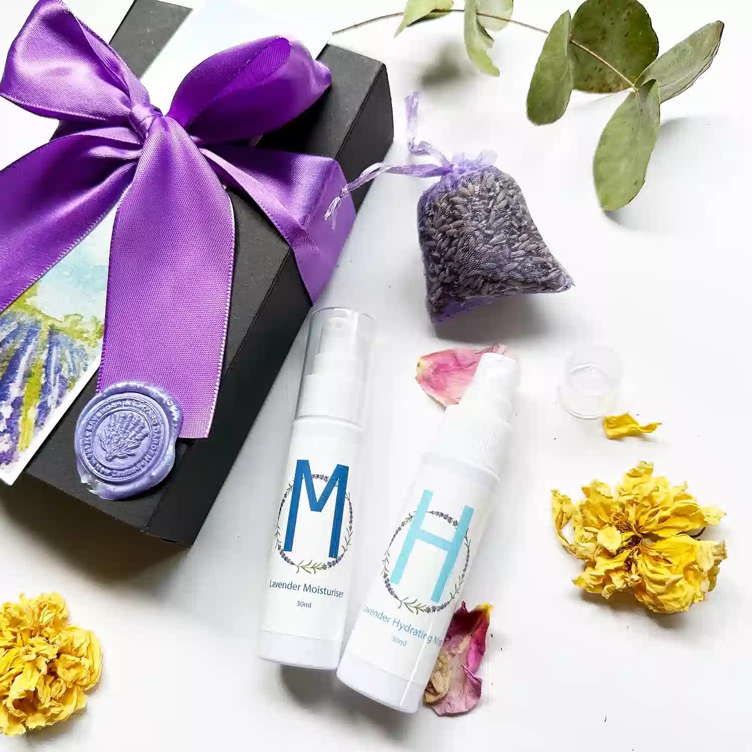 Close-up view of "Moisturizing Lavender Body Lotion, Lavender Hydrosol Floral Water Face Toner, Dried Lavender Sachet" alongside a purple ribbon black gift box with yellow roses and eucalyptus leaves.