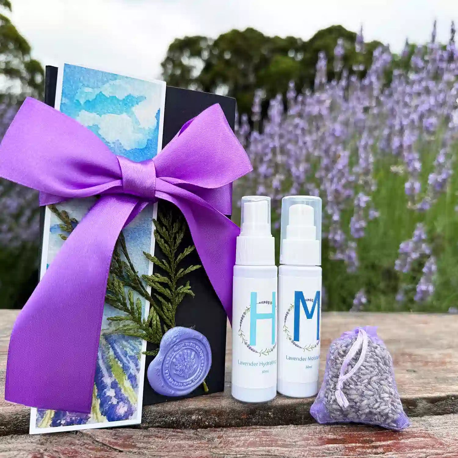 Charming Bridesmaid Presents Set scented by Lavender Essential Oils featuring Moisturizing Lavender Body Lotion, Lavender Hydrosol Floral Water Face Toner, Dried Lavender Sachet, and Lavender Field Bookmark, placed on a wooden bench against purple lavender blooming bushes.