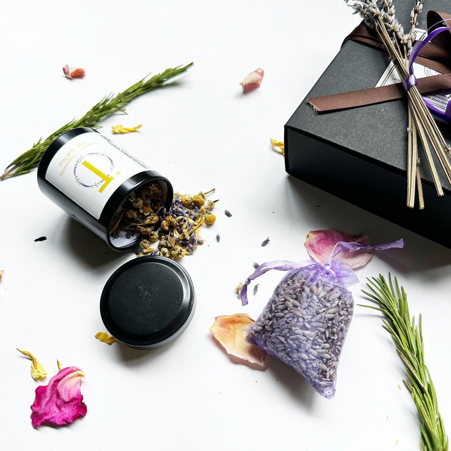 Close-up view of a "Cheap but Cute Gift Ideas for Teacher" set, featuring a black gift box with a brown ribbon. Displayed items include Lavender & Camomile Herbal Tea (Travel Mini) and a Dried Lavender Sachet, surrounded by mini dried lavender flowers, rosemary, and rose petals as background decor.