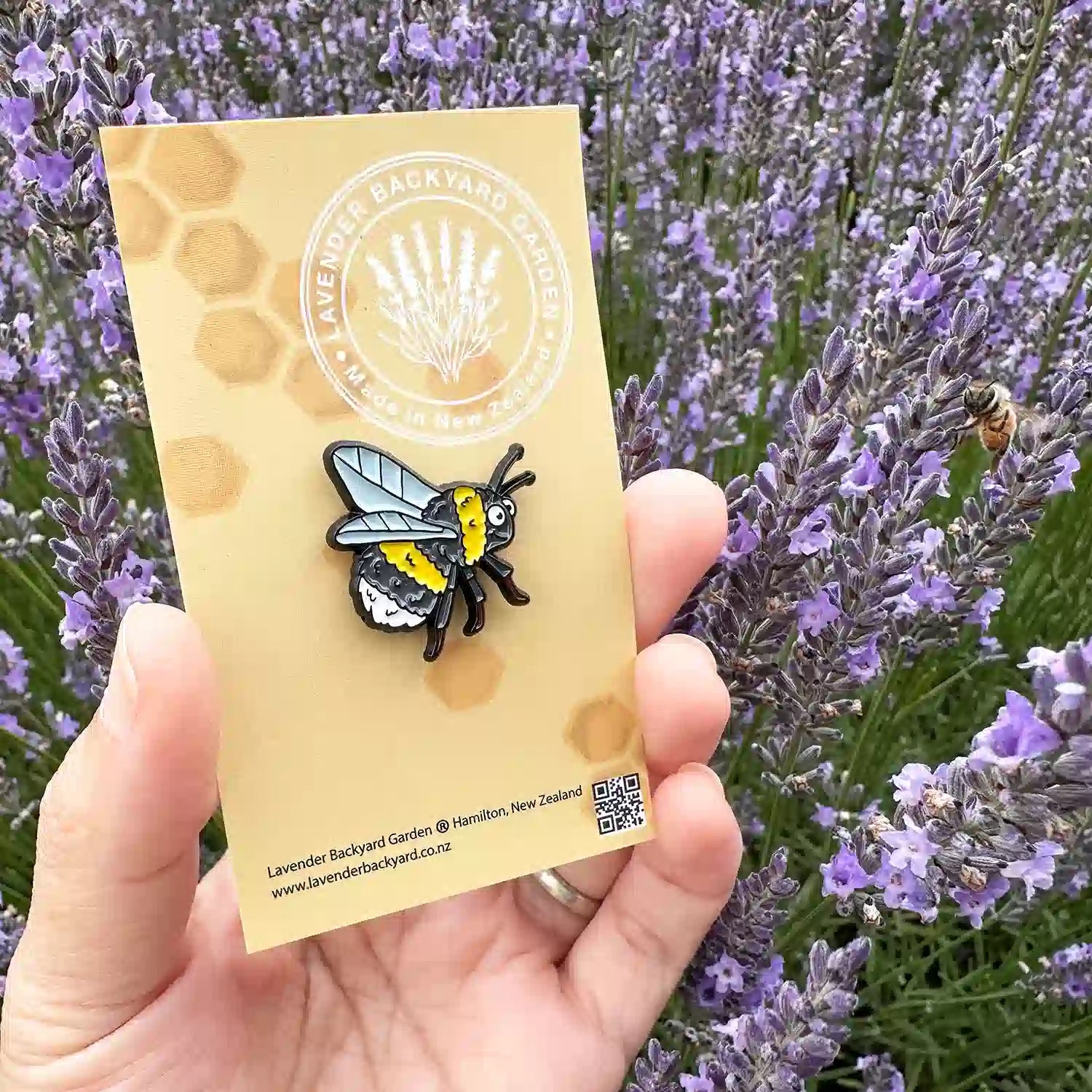 A cute bee enamel badge held in hand against a backdrop of blooming lavender flowers, featuring a real bee buzzing among the lavender.