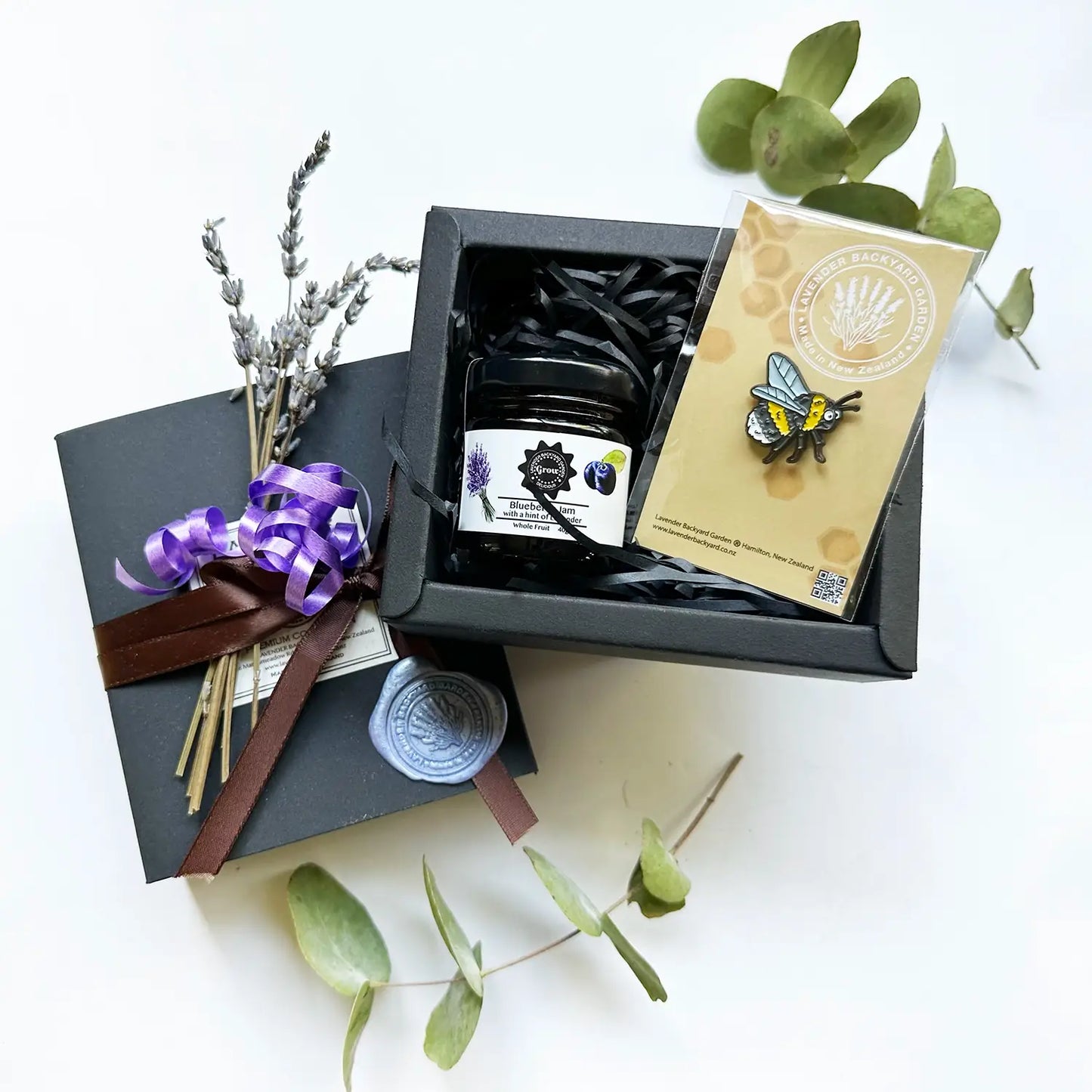 A charming gift box named "Delightful New Zealand Farm Souvenir Gift Set" featuring a cute bee design, containing a New Zealand Lavender Farm souvenir purple tractor badge and lavender blueberry jams, accented with a mini dried lavender flower and wrapped with a brown ribbon.