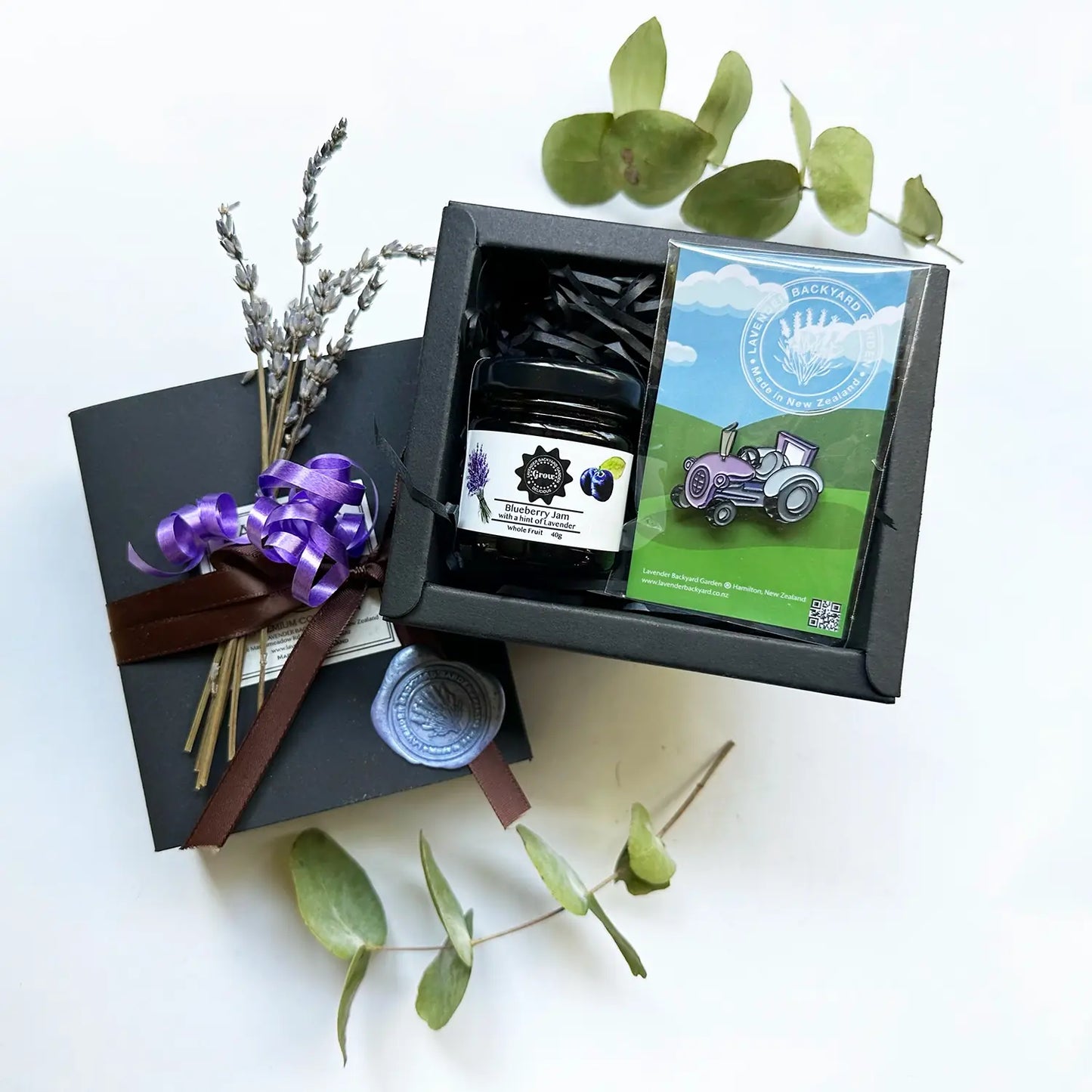 A beautifully arranged gift box featuring a New Zealand Lavender Farm souvenir purple tractor badge, lavender blueberry jams, and a mini dried lavender flower, all wrapped with a charming brown ribbon.