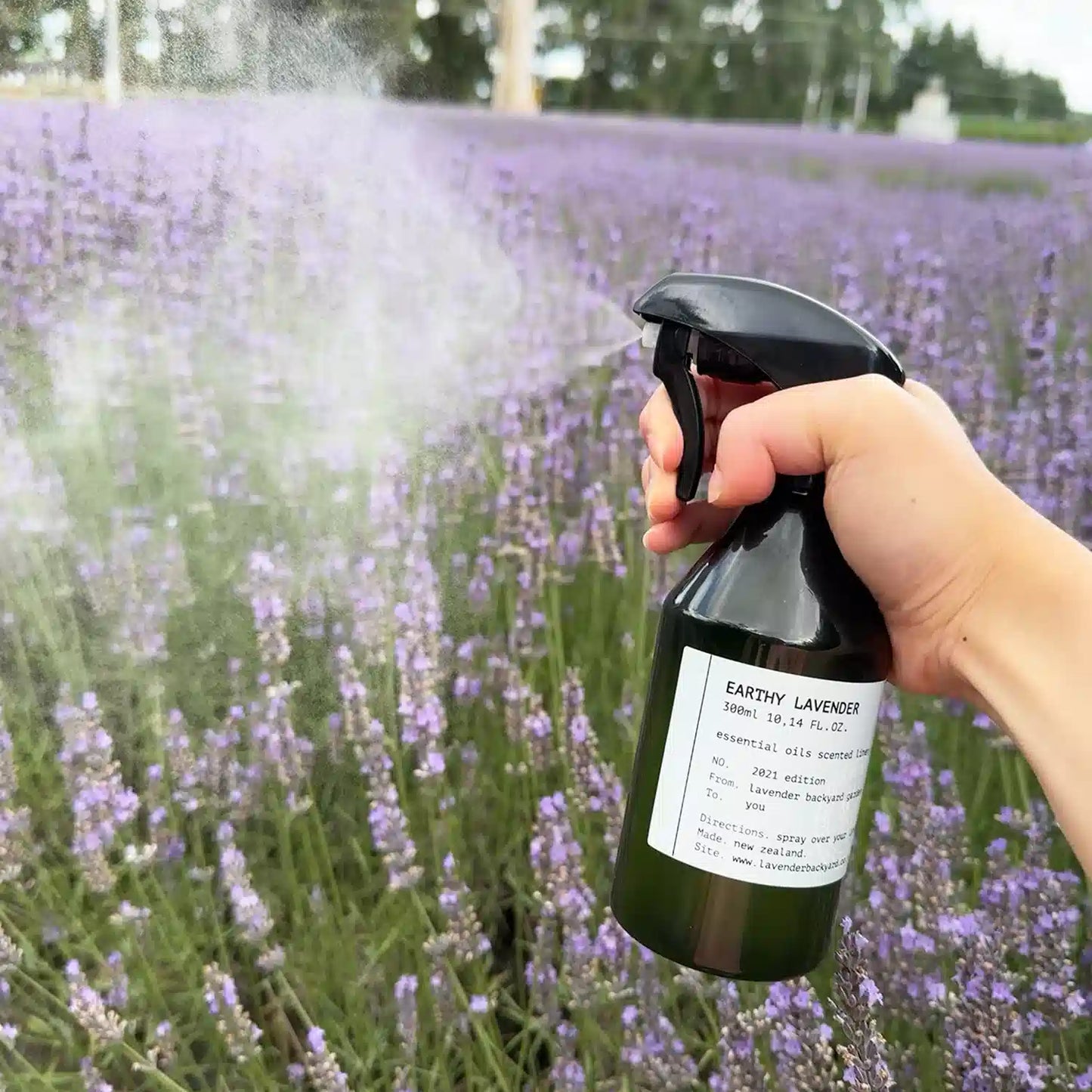 A bottle of earthy lavender linen spray, held by hand and spritzed among a vibrant lavender field at Lavender Backyard Garden in New Zealand.