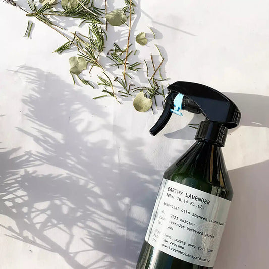 A bottle of earthy lavender linen spray, set against a background of herbs and their shadows used as decoration.