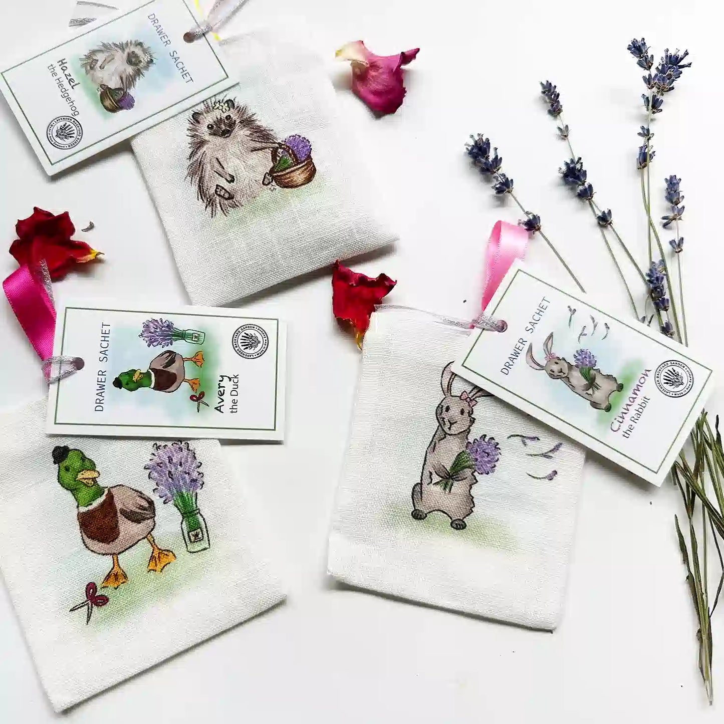 Three farm animal scented linen sachets featuring duck, hedgehog, and rabbit designs, displayed with a background of lavender flowers and rose petal decorations.