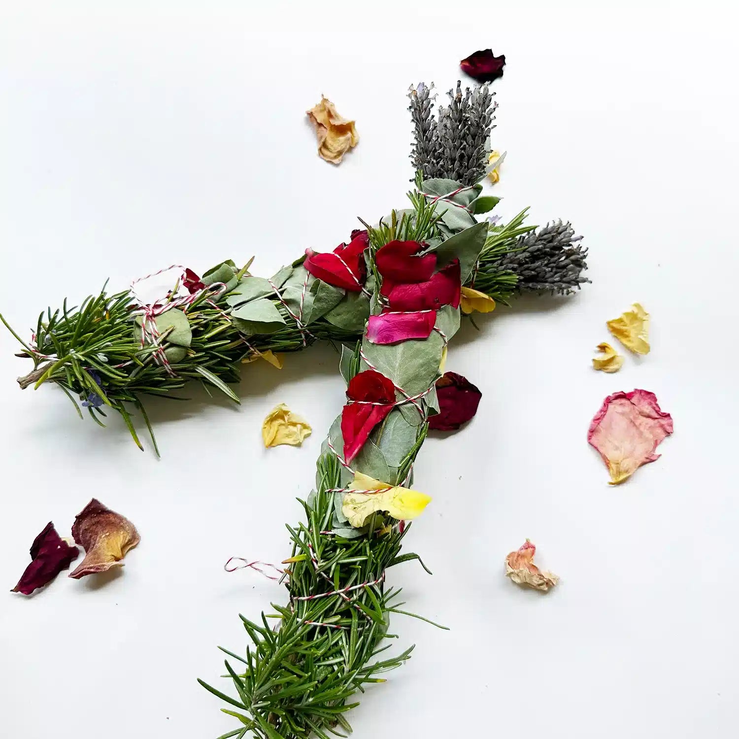 Two freshly made smudge sticks stacked together, containing lavender, cedarwood, white sage, rosemary, eucalyptus, and roses, set against a background of rose petals.