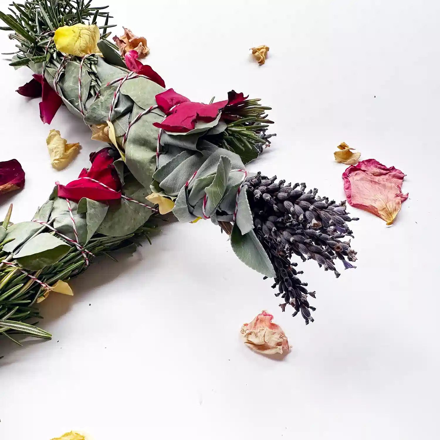 Close-up view of two freshly made smudge sticks containing lavender, cedar-wood, white sage, rosemary, eucalyptus, and roses, set against a background of rose petals.