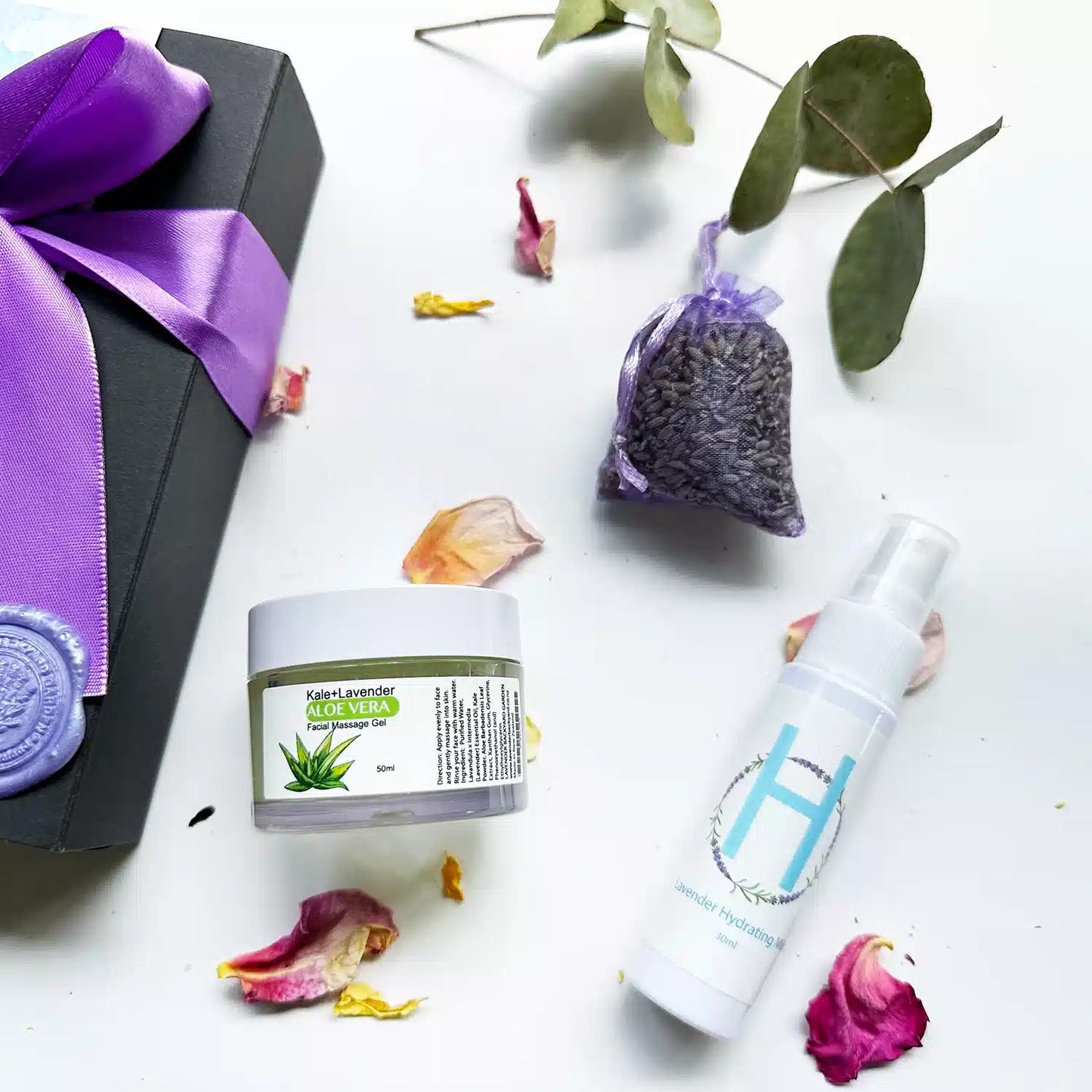 Close-up view of "Glow & Nourish Skin Care Gifts" set, featuring a black gift box with a purple ribbon, surrounded by rose petals and eucalyptus leaves as background decor. Showcased products include a Lavender Hydrosol Floral Water Face Toner (Travel), Dried Lavender Sachet, Lavender Kale Aloe Vera Gel - Exfoliating Facial Massage, and a Lavender Field Bookmark.