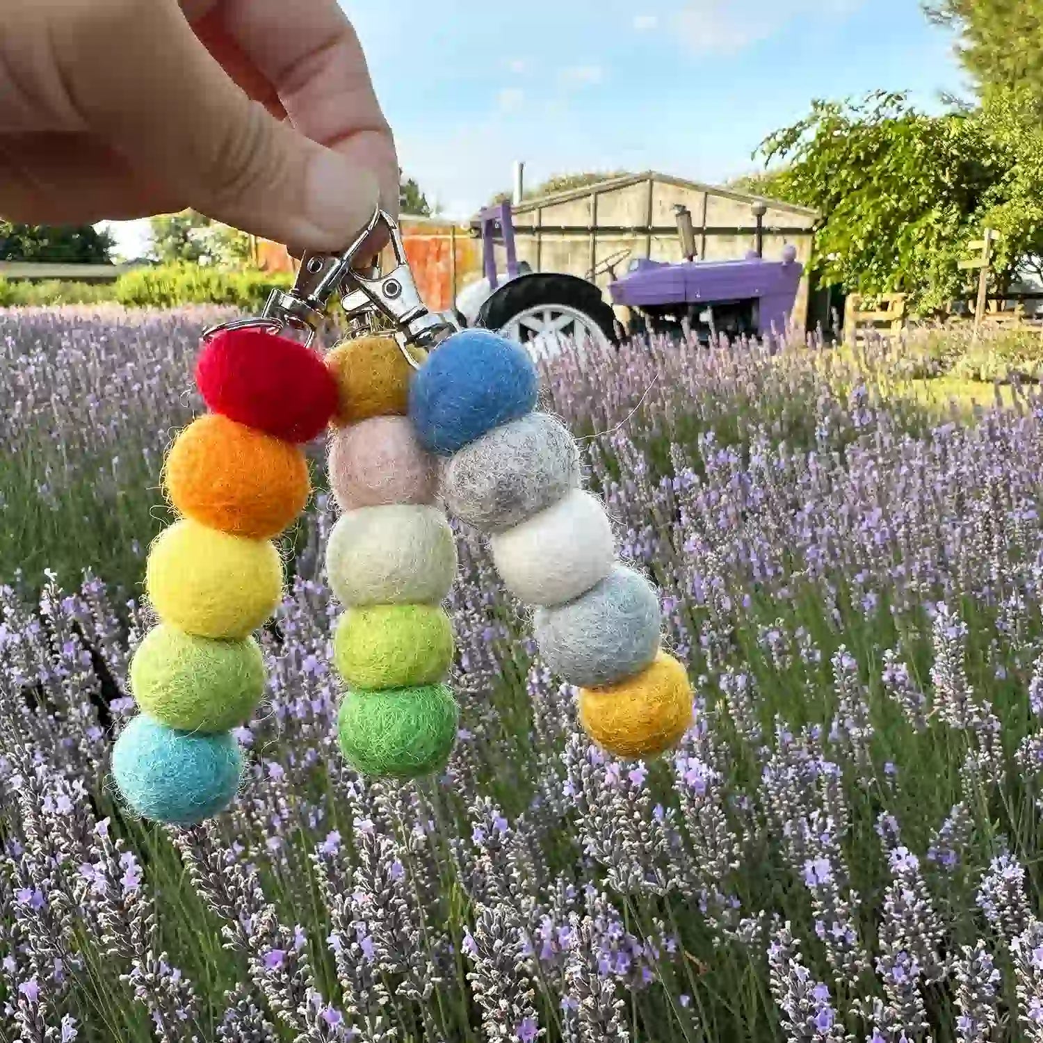 Handcrafted pastel felt ball essential oil keyring diffuser held against a farm shed, tractors, and a lavender purple field at Lavender Backyard Garden NZ.