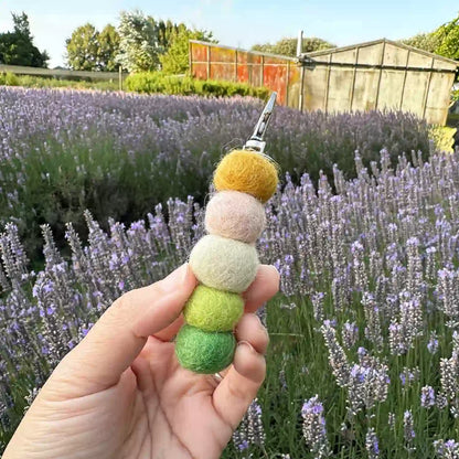 Handmade essential oils felt ball air freshener held against a farm shed with a purple lavender field at Lavender Backyard Garden in New Zealand.