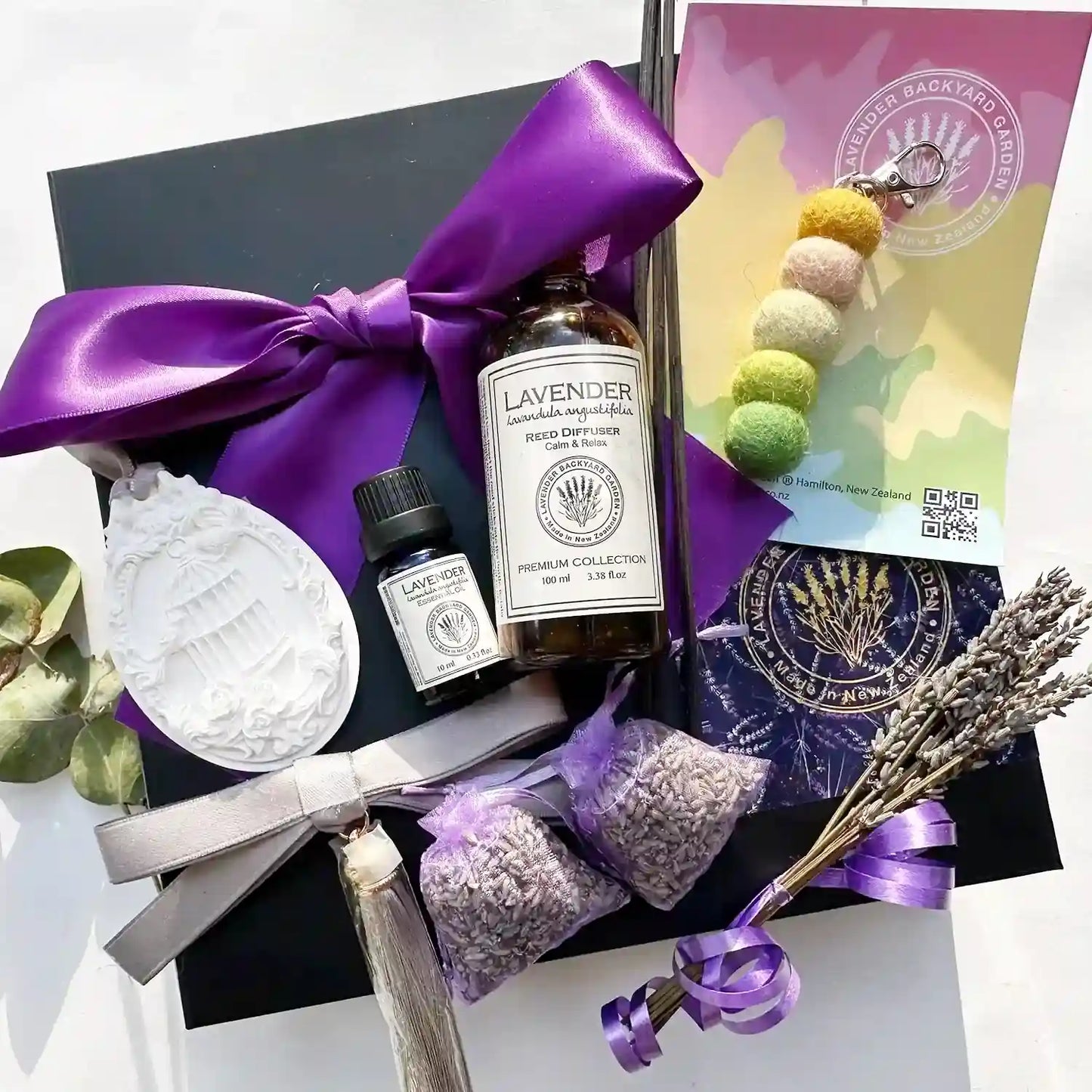 Home Sweet Home Comfort Gift Basket containing a wardrobe aroma stone, lavender essential oil, reed diffuser, and pastel felt ball essential oil keyring diffuser, with a purple ribbon gift box in the background.