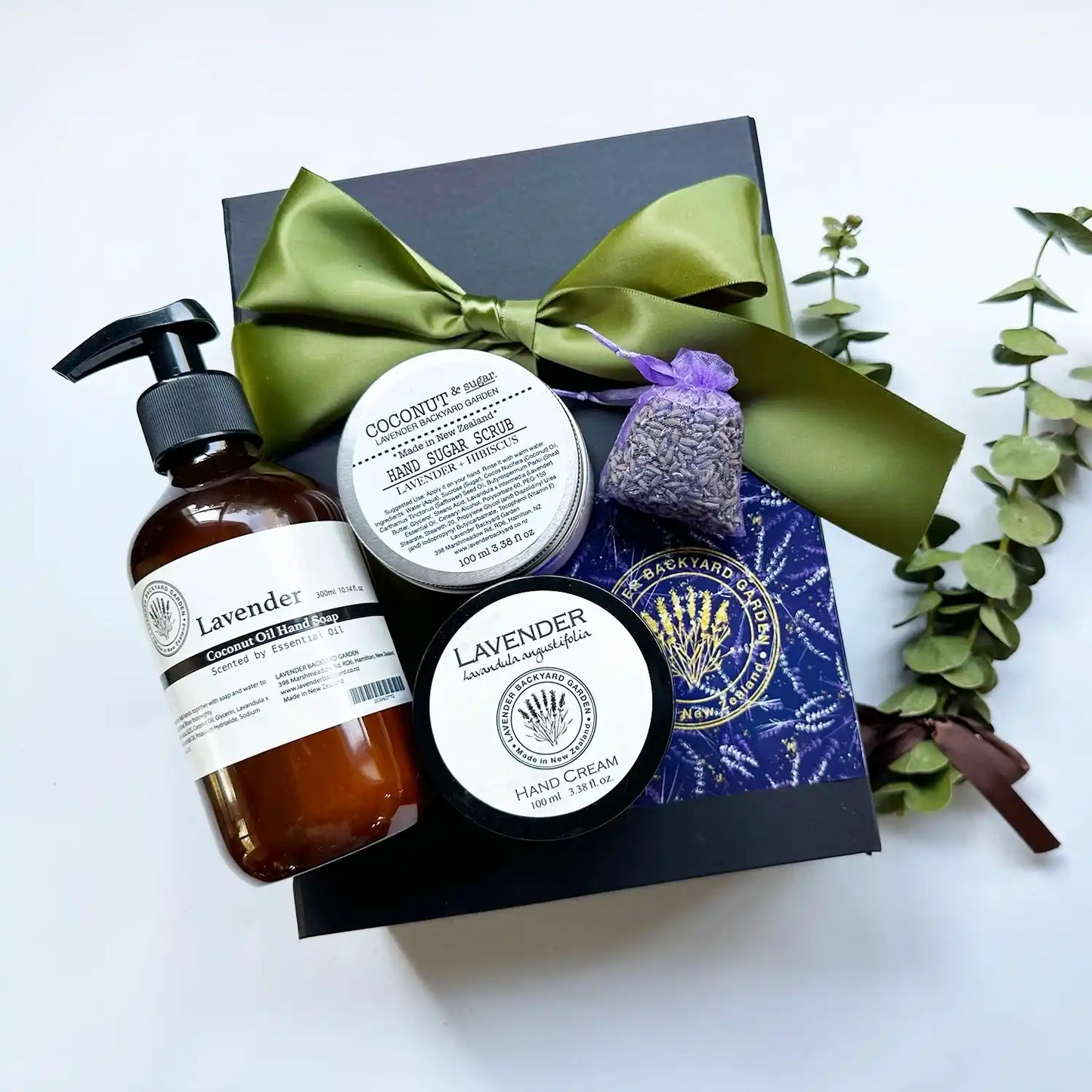 Hydrating liquid hand wash, hand cream, and sugar scrub infused with essential oils, accompanied by a lavender sachet, displayed in a black gift box with a green ribbon background.