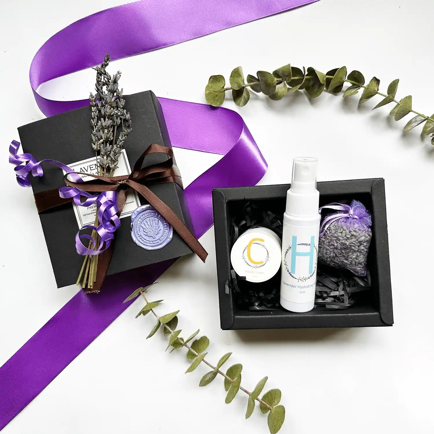 A small gift set featuring travel-size hand cream and facial toner scented with essential oil, elegantly wrapped in a black gift box adorned with a mini dried lavender flower.