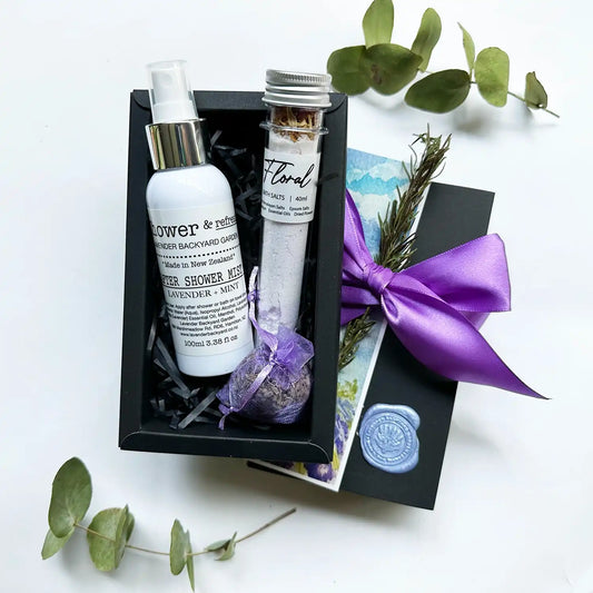 A beautifully wrapped gift box featuring an after shower body mist, lavender bath salt infused with essential oils, and a lavender sachet, elegantly tied with a purple ribbon and accompanied by a lavender field bookmark.