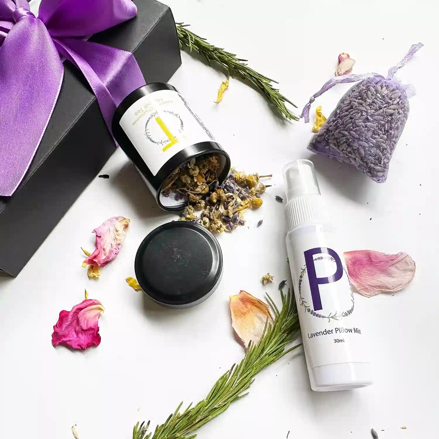 Close-up view of the "Indulgent Relaxing Gift Set for Mother," featuring Lavender Sleep Spray (Travel Size), Lavender & Camomile Herbal Tea - Loose Leaf (Travel Size) with the open lid revealing chamomile and lavender dried flowers, and a Dried Lavender Sachet. The background includes rosemary, rose petals, and a black gift box with a purple ribbon.