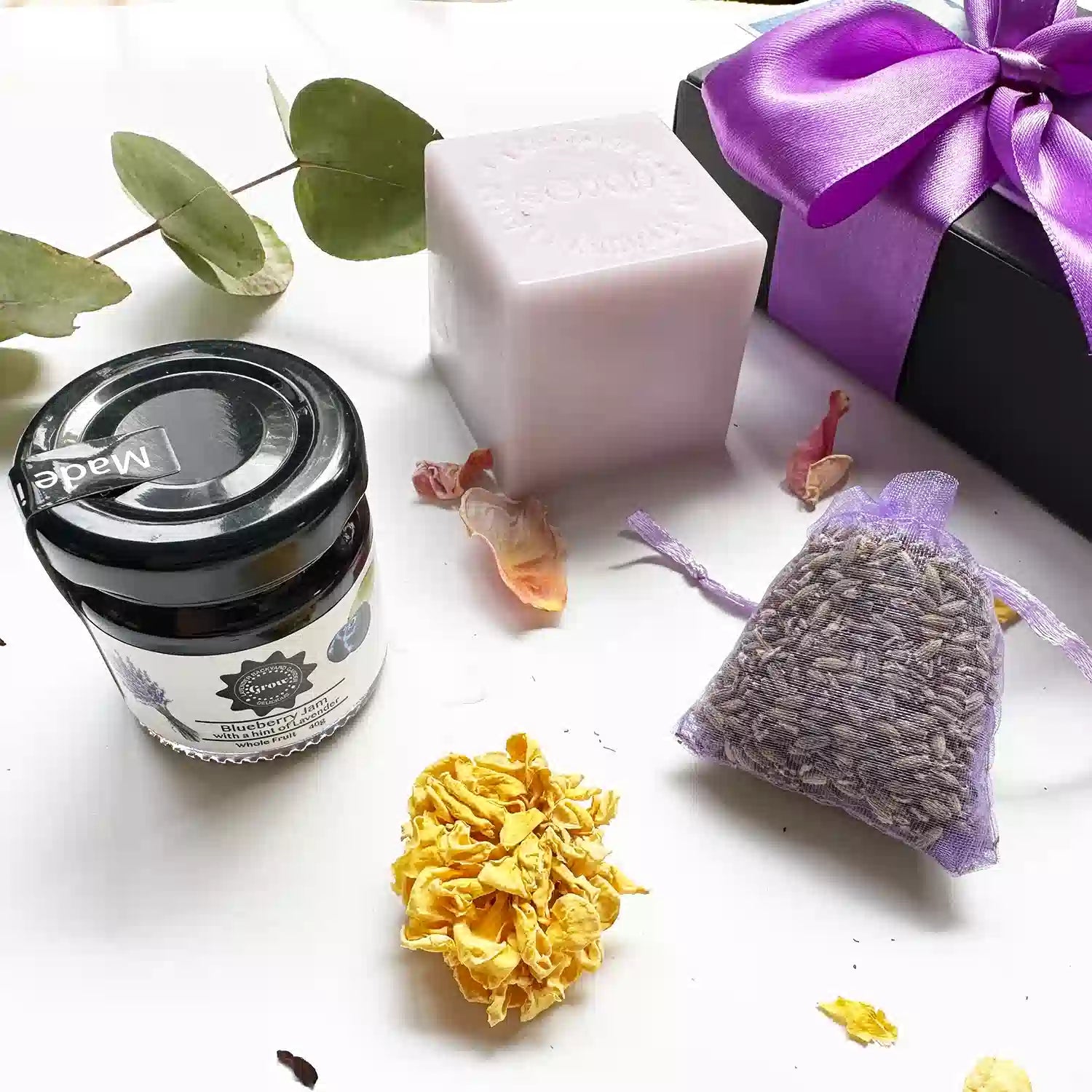 Close-up view of an "Inexpensive yet Thoughtful Gift Box," featuring a black gift box with a brown ribbon. Showcased items include a Dried Lavender Sachet, Blueberry Jam with a hint of Lavender (40g), and Lavender Goat Milk Handmade Soap, surrounded by mini dried lavender flowers, rosemary, and rose petals as background decor.