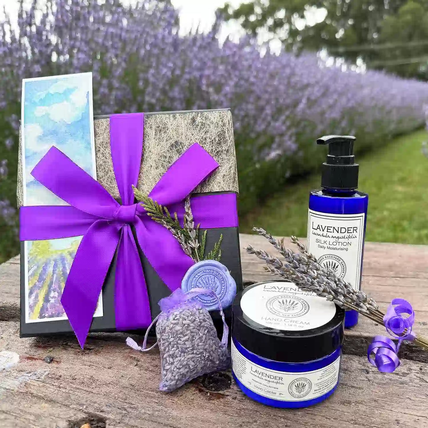 Lavender Beauty Skincare Gift Set scented by essential oils, displayed on a wooden bench against a purple blooming lavender field, featuring products Lavender Hand Cream, Lavender Silk Lotion, Dried Lavender Sachet, Mini Lavender Dry Flower, and Lavender Field Bookmark.