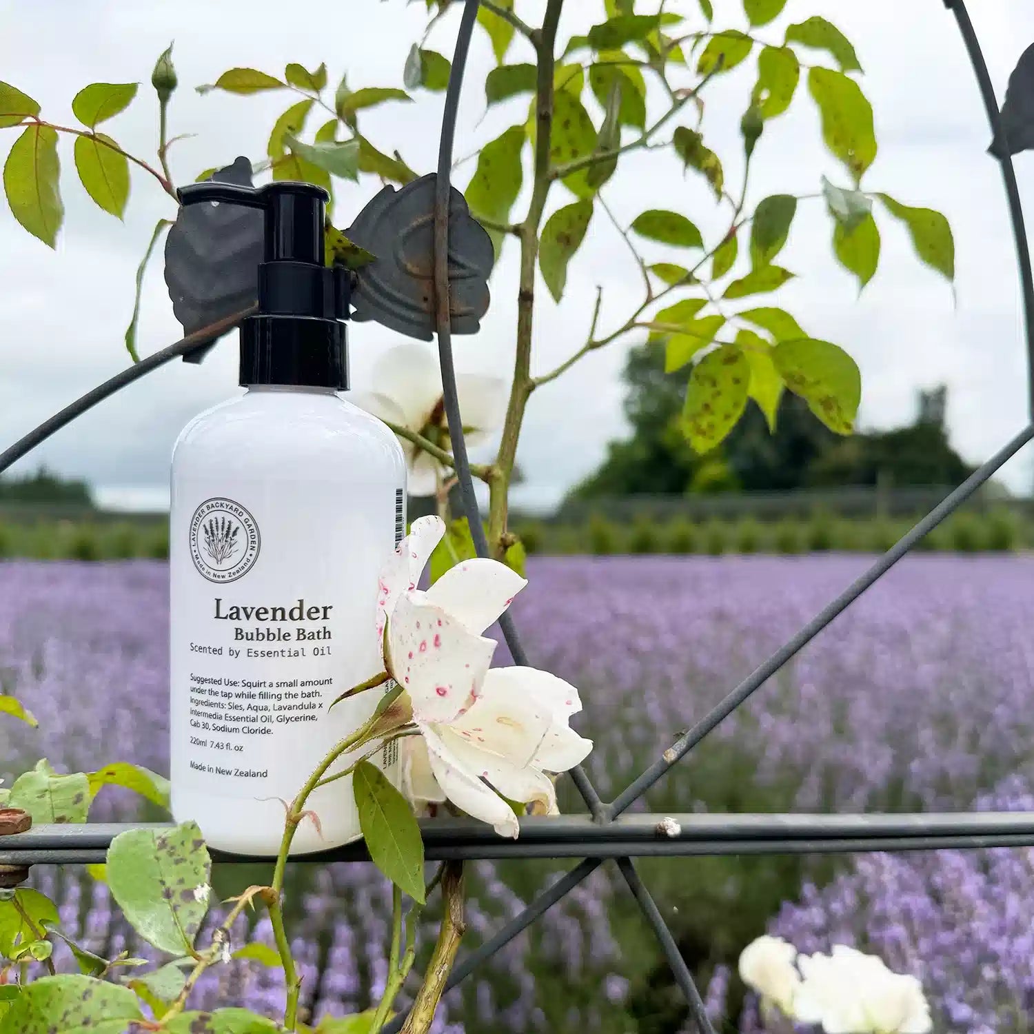 A bottle of lavender bubble bath scented with essential oil, made in New Zealand, set against a calming background of soft lavender hues and a white rose.