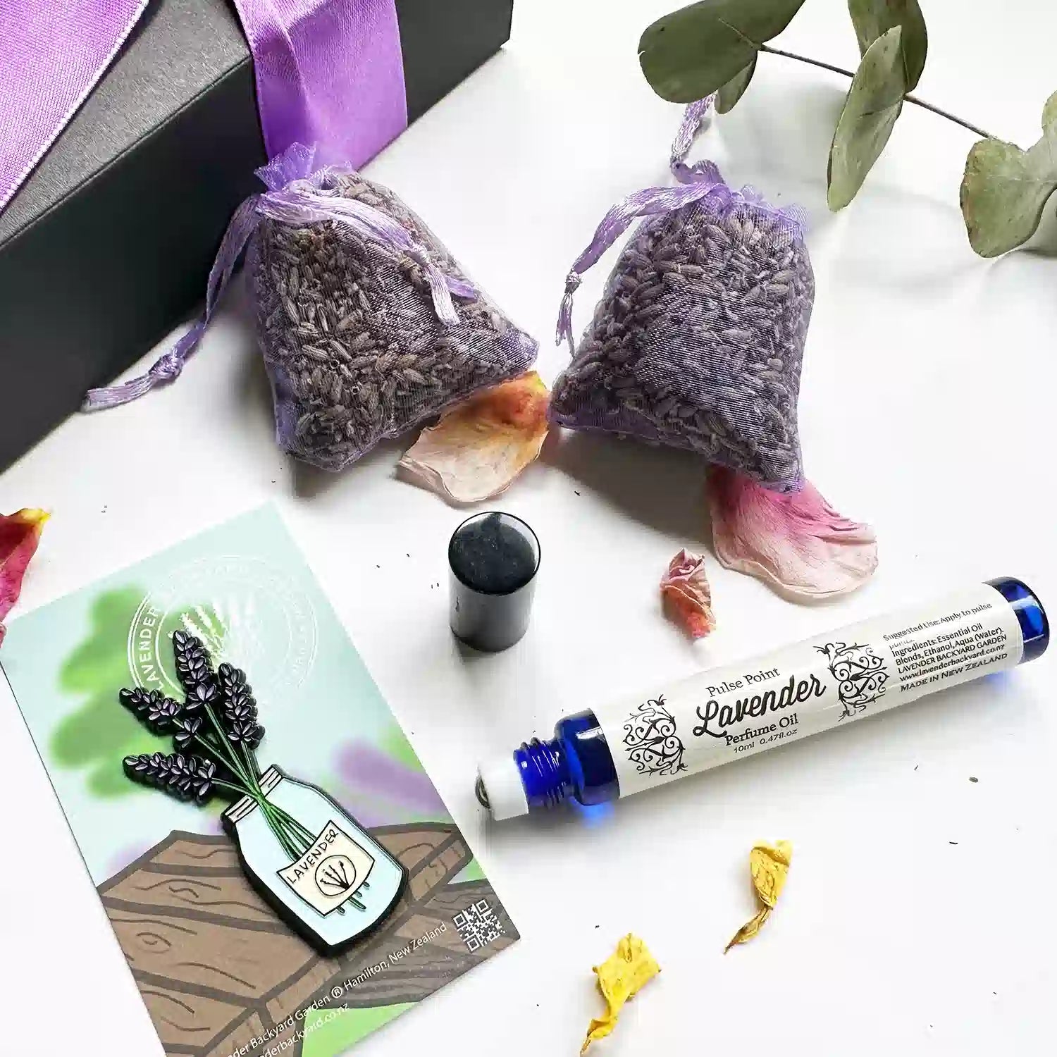 Close-up view of "Natural Light Perfume Pulse Point, Jar of Lavender Fridge Magnets, and 2 Dried Lavender Drawer Sachets," decorated with eucalyptus leaves and rose petals, with a purple ribbon gift box in the background.