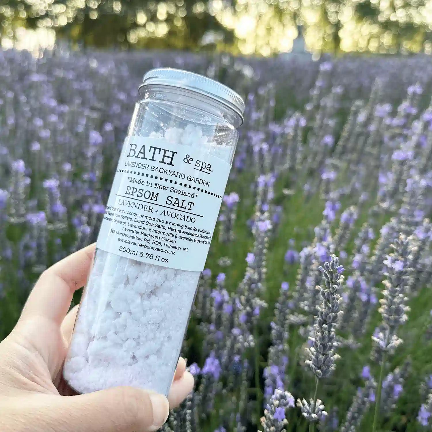 Lavender Epsom Bath Salts held by hand against a backdrop of purple blooming lavender at Lavender Backyard Garden, NZ.