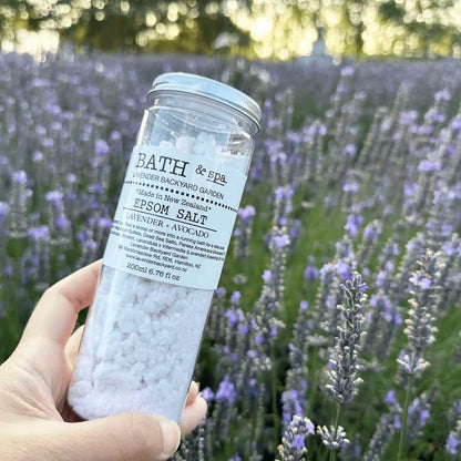 Lavender Epsom Bath Salts held by hand against a backdrop of purple blooming lavender at Lavender Backyard Garden, NZ.