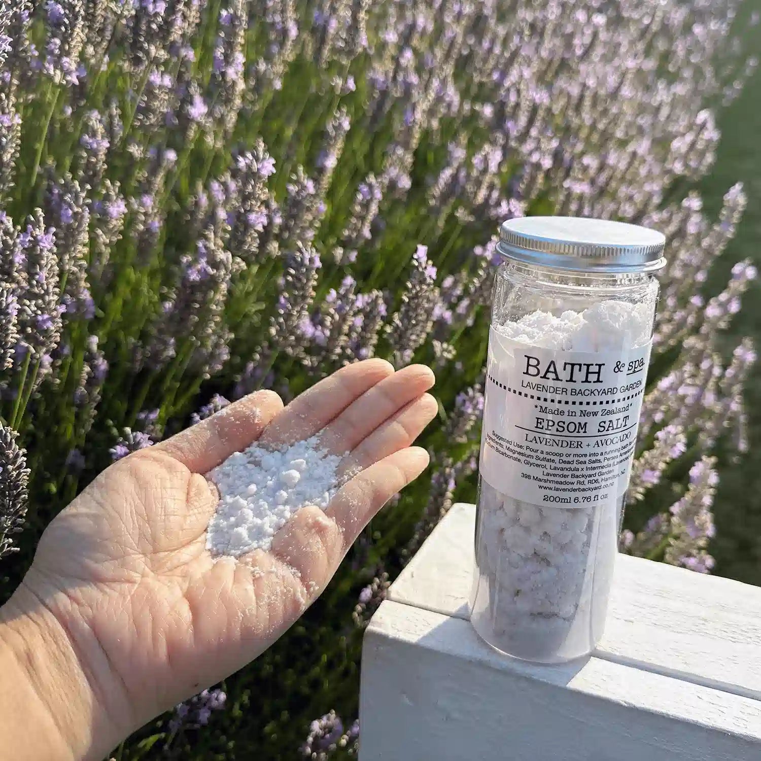 A bottle of lavender epsom salt placed on a white wooden chair, one hand holding the salts to show the texture, against a purple sunset lavender field in New Zealand.