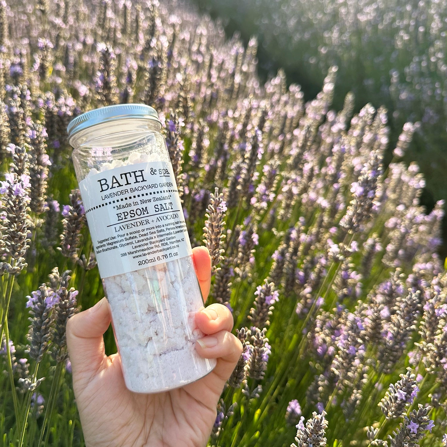 Lavender Epsom Bath Salts held by hand against a backdrop of purple blooming lavender at Lavender Backyard Garden, NZ, during sunset.