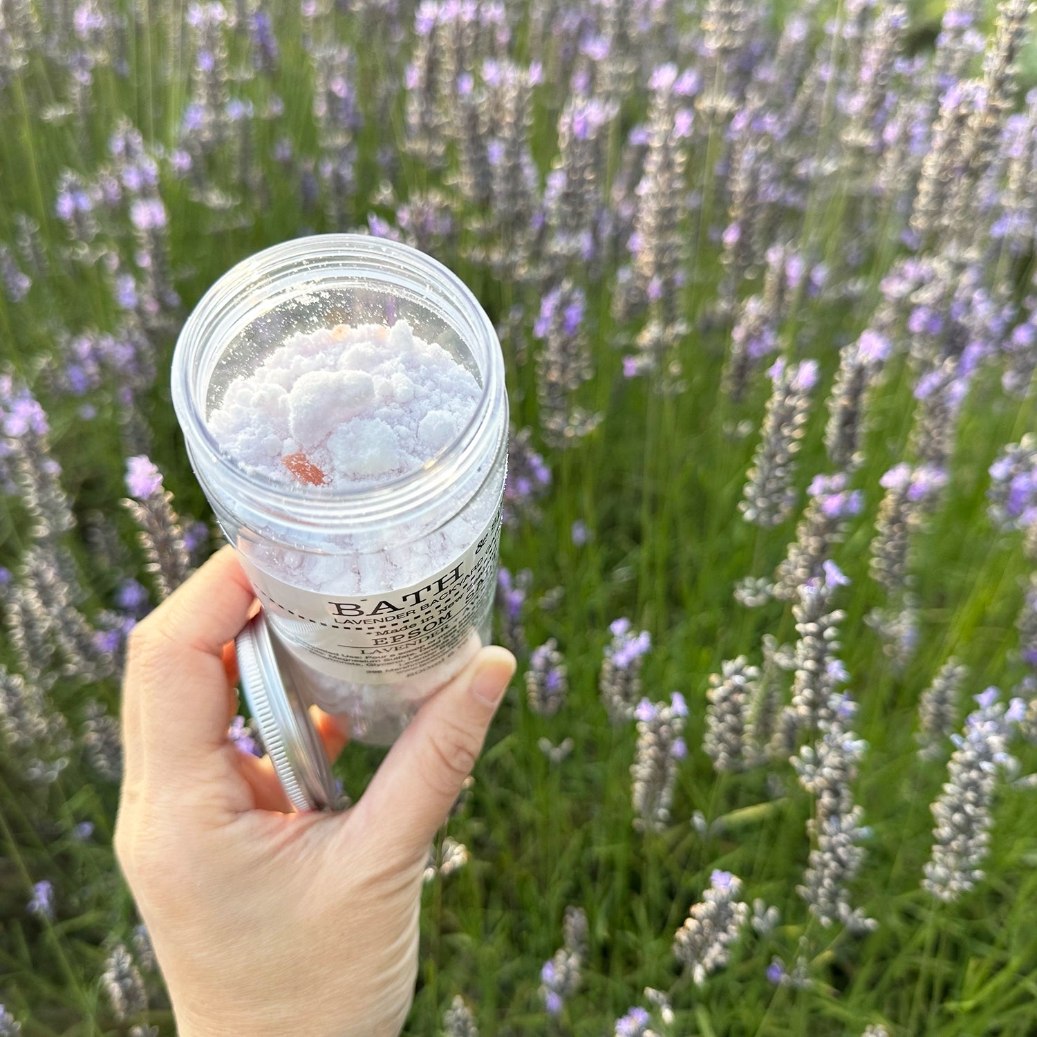 Open lid of Lavender Epsom Bath Salts bottle, showcasing the texture of the Epsom salt inside, held by hand against a backdrop of purple blooming lavender at Lavender Backyard Garden, NZ.