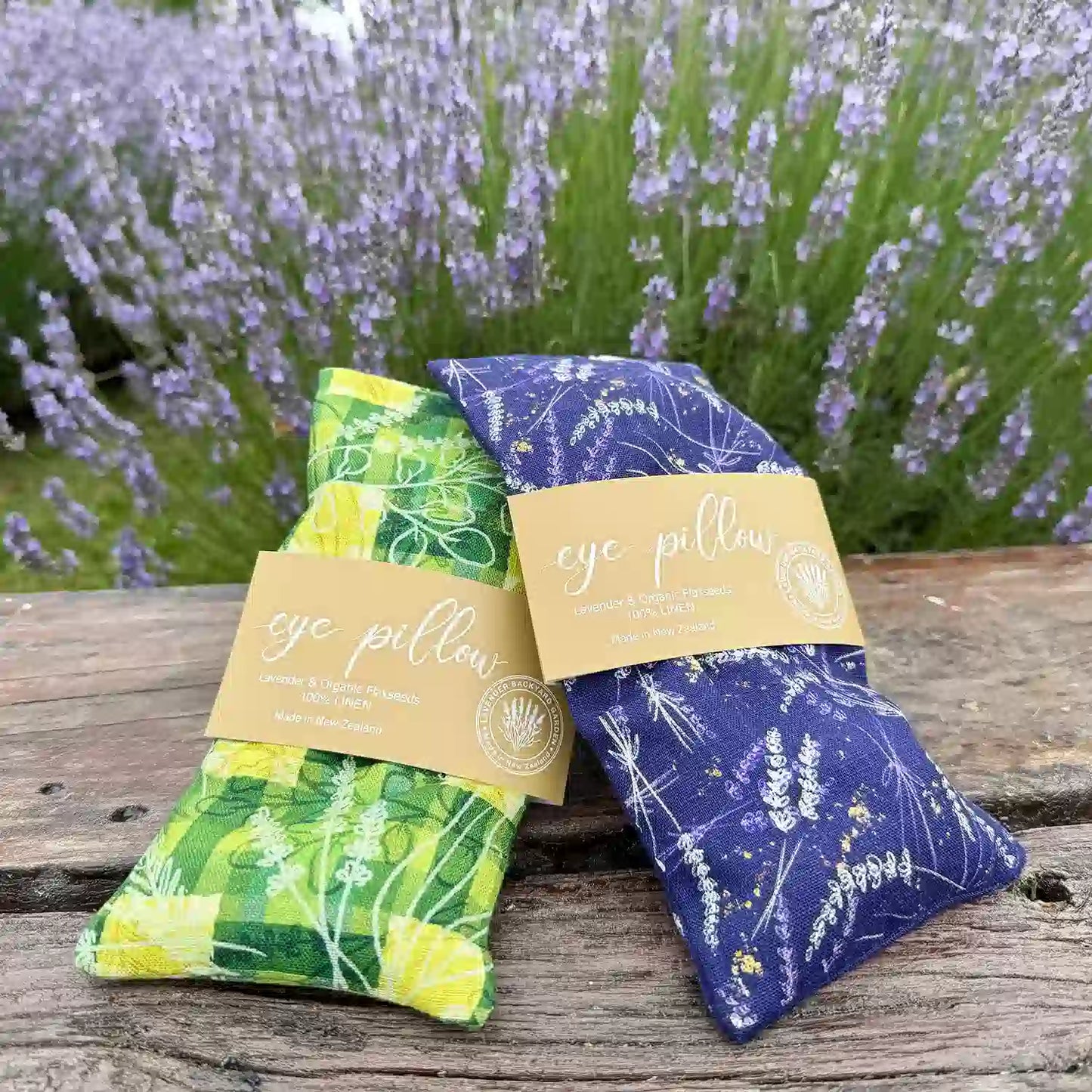 Two lavender eye pillows, one with a green pattern and the other with a purple pattern, placed on a bench against blooming lavender plants.