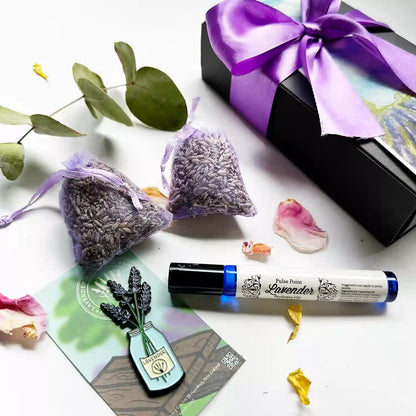 Close-up view of "Natural Light Perfume Pulse Point, Jar of Lavender Fridge Magnets, and 2 Dried Lavender Drawer Sachets" decorated with eucalyptus leaves and rose petals, with a purple ribbon gift box in the background.