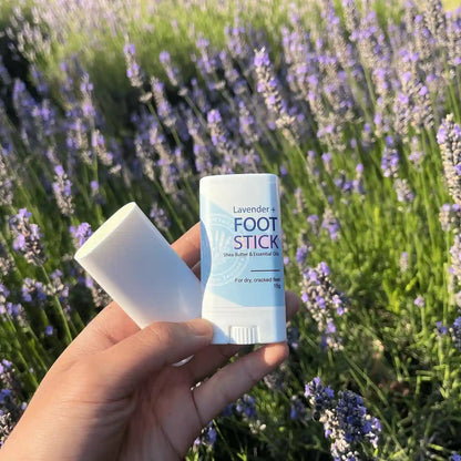 One lavender foot balm for caring feet, scented by lavender essential oils, held by hand against blooming lavender bushes. The lid is open, showcasing the balm's texture.