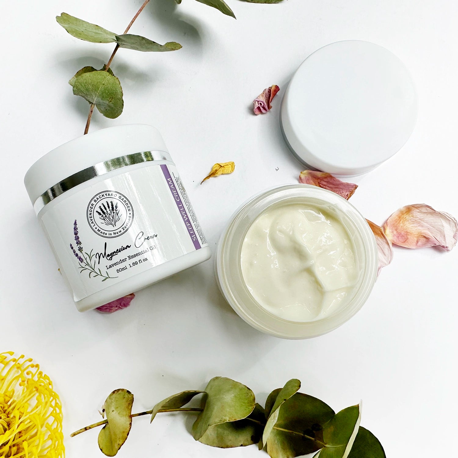 Close-up view of Lavender Magnesium Sleep Cream with two pots; one with the lid off, showing the smooth texture of the cream, with eucalyptus leaves, yellow flowers, and rose petals in the background.