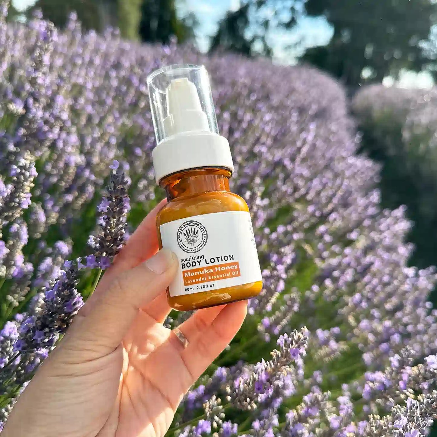 Lavender Manuka Honey Body Moisturizer, held by hand against purple blooming lavender rows at Lavender Backyard Garden, New Zealand.