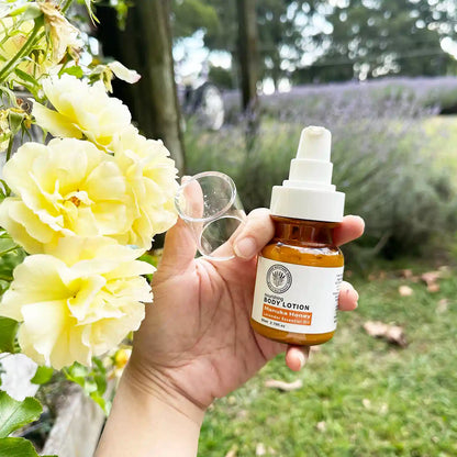 Lavender Manuka Honey Body Moisturizer, held by hand with open lid, placed beside yellow roses against a purple lavender field at Lavender Backyard Garden, New Zealand.