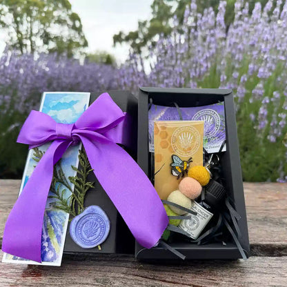 Lavender Missing You Joyful Cute Gift Box scented by lavender essential oils, placed on a wooden bench against purple blooming lavender fields at Lavender Backyard Garden, NZ, featuring products Aromatherapy Pure Lavender Essential Oils, Pastel Felt Ball Keyring Car Diffuser, Very Cute Bee Badge, and Lavender Field Bookmark.