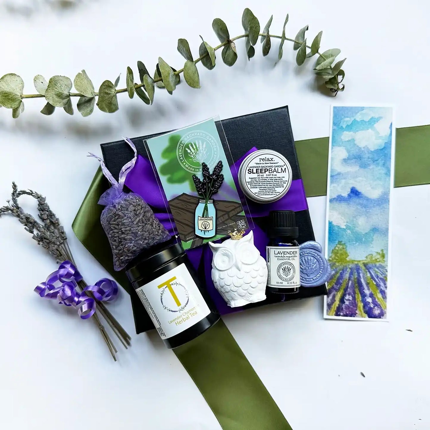 A beautifully crafted Lavender Pamper Gift Set for Mum featuring a lavender dried sachet, sleep balm, lavender essential oil, owl aroma stone, lavender flower fridge magnet, and lavender chamomile tea, all wrapped elegantly with a purple ribbon in a black gift box with a lavender field bookmark.