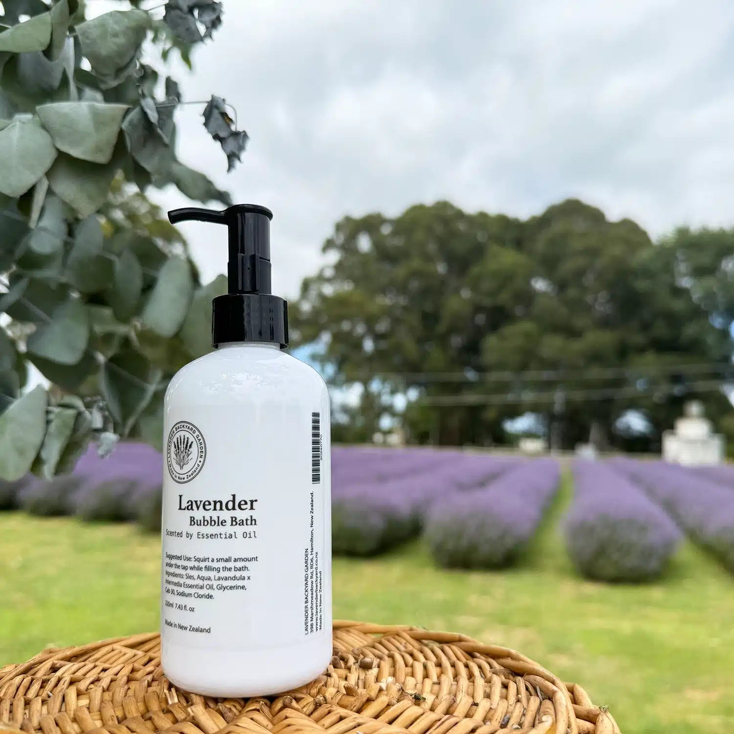 A bottle of lavender-scented bubble bath made in New Zealand, placed on a basket, with the vibrant lavender field at Lavender Backyard Garden in New Zealand in the background.